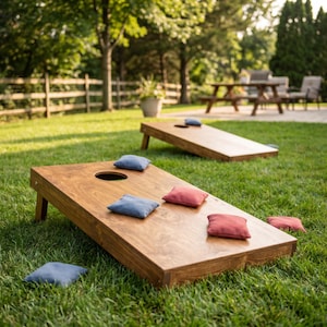 Könnte beinhalten: Zwei rechteckige Cornhole-Spielbretter aus Holz mit kreisförmigen Löchern, die auf einem Rasen stehen. Mehrere quadratische Sitzsäcke in Blau- und Rottönen sind auf den Brettern und dem Gras verteilt, die für das Wurfspiel verwendet werden.