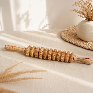 May include: A wooden massage roller tool featuring a long handle on each side and a series of ten rotating, notched circular discs in the center. The wood displays a natural grain pattern with light brown, tan, and subtle reddish-orange swirls.