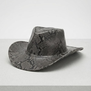 Puede incluir: Un sombrero de vaquero con un patrón texturizado de piel de serpiente en gris y negro. El sombrero tiene una copa pellizcada y un ala ancha y curva, con una banda de piel de serpiente a juego que rodea la base de la copa, creando un aspecto detallado.