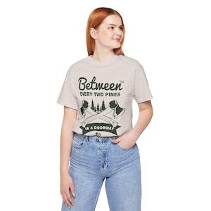 May include: A light gray short-sleeve t-shirt featuring a dark green graphic of two crossed axes, pine trees, and the text "Between every two pines is a doorway to a new world." The design is centered on the chest of the crew neck garment.