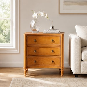 May include: A light brown wooden chest of drawers with three curved front drawers, each featuring two dark metal ring pulls. On top sits a clear glass vase with white orchids, a silver bell, and two books titled The Art of Interiors and Form and Light.