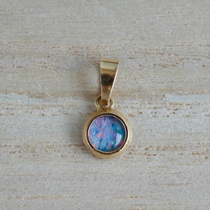 Pode incluir: Um pingente de ouro com uma pedra cabochão redonda em cravação inglesa, apresentando um jogo de cores iridescentes em azul e rosa. O pingente inclui uma argola de ouro polido no topo, projetada para fixação em uma corrente ou cordão.
