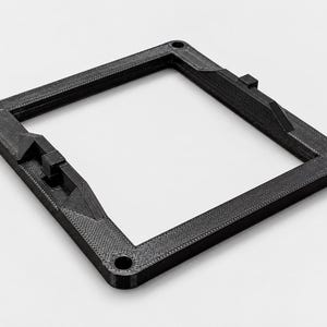 May include: A black, square-shaped 3D printed frame with a textured surface. The frame features four circular mounting holes at each corner and two raised, wedge-shaped brackets with small rectangular tabs positioned on opposite inner sides.