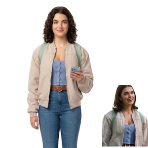 May include: A beige bomber jacket worn over a light blue button-down crop top, paired with blue denim jeans featuring a brown leather belt with a circular buckle. A light green backpack is visible on the shoulders, and a smartphone is held in hand.