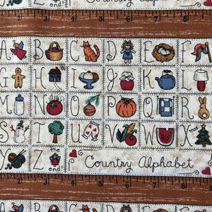 May include: A fabric print featuring a grid of letters from A to Z, each paired with a small illustration like a cat, pumpkin, or snowman. The design includes a brown border with a ruler pattern marked in inches and the text Country Alphabet.
