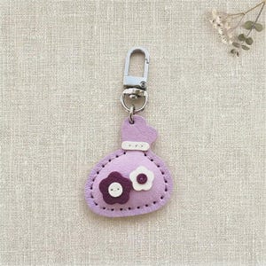 Könnte beinhalten: Ein lavendelfarbener Schlüsselanhänger aus Leder in Form eines kleinen Beutels mit einem silberfarbenen Karabinerhaken. Der Beutel hat ein weißes Zierband oben und zwei kleine blumenförmige Applikationen in Dunkellila und Weiß mit je einem Knopf.