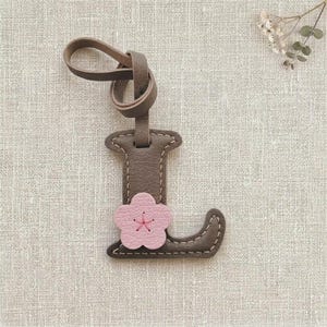 Puede incluir: Un llavero de cuero color topo con forma de letra L y costuras claras visibles en los bordes. Una pequeña flor de color rosa claro de cinco pétalos con un centro cosido en rosa oscuro está sujeta a la parte frontal de la letra.