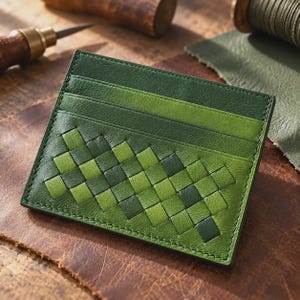 May include: A rectangular leather card holder featuring a woven pattern of alternating dark and light green strips. The top section consists of two horizontal solid green leather slots for cards, with visible stitching along the edges of the item.