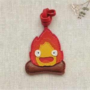 Puede incluir: Un llavero de cuero con forma de llama en rojo y naranja que presenta una cara amarilla con dos ojos blancos y una boca ancha, sobre una base de tronco marrón. El diseño incluye un lazo de cuero rojo en la parte superior con costuras visibles.