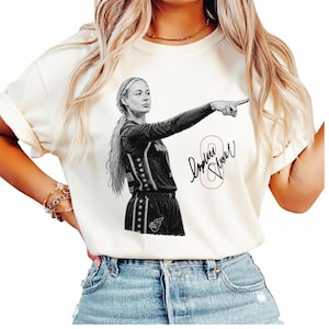 May include: A cream-colored short-sleeve t-shirt featuring a black and white graphic illustration of a basketball player pointing forward. The design includes a stylized signature and the number 8 in a thin red outline next to the player figure.