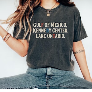 May include: A charcoal grey short-sleeve t-shirt featuring the text GULF OF MEXICO, KENNEDY CENTER, LAKE ONTARIO. The letters F, D, and T are highlighted in red and blue, while the remaining text is in a cream-colored serif font.