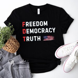 May include: A black short-sleeved t-shirt featuring the white text FREEDOM, DEMOCRACY, and TRUTH stacked vertically. The letters F, D, and T are highlighted in red. A small, distressed American flag graphic is positioned next to the word TRUTH.