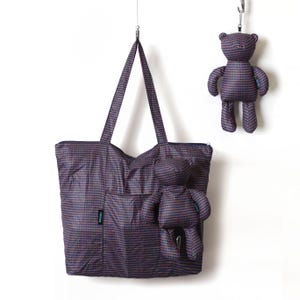 Pode incluir: Uma sacola e um chaveiro de urso de pelúcia, ambos em tecido roxo escuro com um padrão repetido de pequenos pontos azuis e vermelhos. A sacola tem um bolso frontal e fecho de zíper. O chaveiro de urso de pelúcia está pendurado em um gancho de metal.