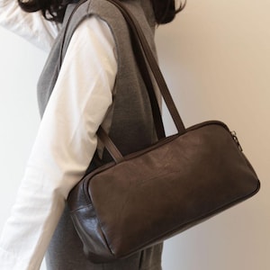 May include: A brown rectangular handbag with a zipper closure and two handles. The bag is made of a textured material and is being carried over the shoulder. The bag is a dark brown color.