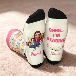 May include: A pair of cream-colored socks with black heels and toes. One sock features a cartoon of a person reading with books, while the other displays the text SHHH... I'M READING in pink, along with a book icon and the placeholder text Name.