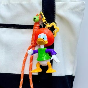 May include: A white and black canvas tote bag featuring a hanging keychain accessory. The keychain includes a plastic Donald Duck figure with an orange afro wig, a green shirt, purple sleeves, and yellow feet, attached to an orange paracord lanyard.