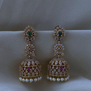Peut inclure: Une paire de boucles d'oreilles jhumka dorées avec un design en forme de cloche orné de cristaux transparents, une pierre verte en forme de goutte en haut, une pierre rose sur la cloche et une rangée de petites perles blanches en bas.