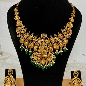 May include: A gold-toned necklace and matching pair of earrings featuring intricate embossed deity figures, ornate peacock motifs, and dangling green beads accented with small white pearls, displayed on a black velvet jewelry stand.