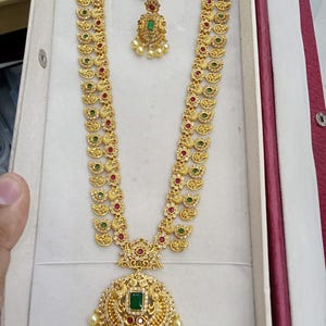 May include: A gold-toned long necklace and matching pair of earrings featuring intricate metalwork, small green and red stones, and white pearl-like beads. The necklace has a large circular pendant with a central green stone and dangling beads.