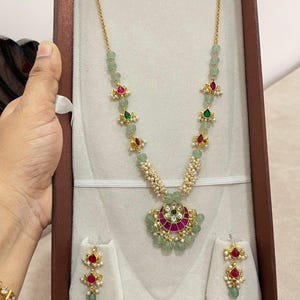 Puede incluir: Un conjunto de collar y pendientes en tono dorado con cuentas verde claro, pequeñas perlas blancas y detalles en forma de lágrima rojos y verdes. El collar incluye un colgante central en forma de media luna con detalles florales.