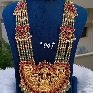 Puede incluir: Un conjunto de collar y pendientes en tono dorado con un gran colgante central con motivos de deidades, acentuado por piedras rojas y verdes. El collar consta de múltiples hileras de pequeñas cuentas verdes, intercaladas con piezas doradas.