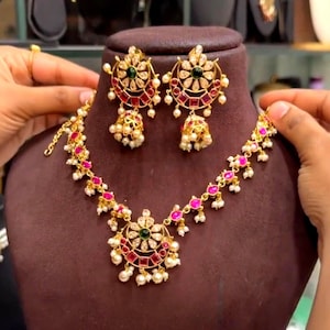 Könnte beinhalten: Ein goldfarbenes Schmuckset bestehend aus einer Halskette und passenden Jhumka-Ohrringen. Jedes Teil zeigt ein halbmondförmiges Design mit klaren Steinen, rechteckigen roten Steinen, einem grünen Mittelstein und hängenden weißen Perlen.