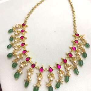 May include: A gold-tone necklace featuring a series of teardrop-shaped fuchsia stones interspersed with small white pearls and dangling green oval beads. The chain consists of circular links, and the pendant section displays a symmetrical arrangement.