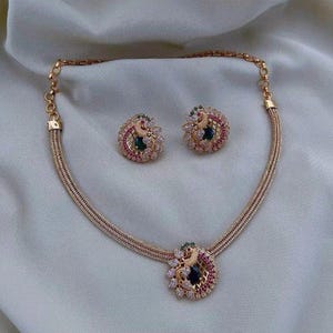 Peut inclure: Un ensemble collier et boucles d'oreilles assorties en ton doré avec un motif de paon. Chaque pièce est ornée de cristaux blancs, de petites pierres roses et d'une pierre ovale bleu foncé centrale, serties dans un treillis métallique.