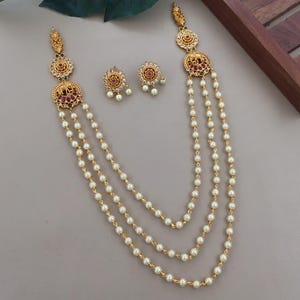 May include: A gold-toned necklace and earring set featuring three strands of white pearls. The necklace includes ornate circular pendants with elephant motifs, red and green stones, and clear crystals. Matching stud earrings feature a similar circular design.