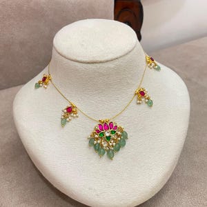 Puede incluir: Un collar de tono dorado con un colgante central en forma de flor de loto con piedras rosas y verdes, acentuado por pequeñas perlas blancas. La cadena incluye dijes adicionales con forma de flor con piedras rosas y cuentas verdes en gota.