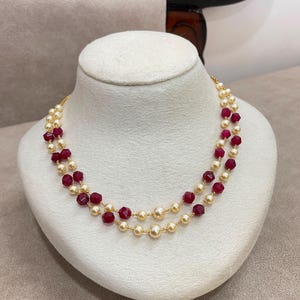 Puede incluir: Un collar de doble hebra que presenta perlas esféricas de color crema alternadas con cuentas facetadas de color rojo oscuro. Las cuentas están ensartadas en una fina cadena de tono dorado, creando una apariencia en capas con un patrón constante.