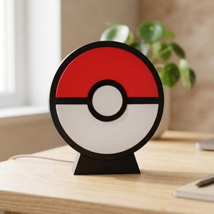May include: A circular lamp shaped like a Poké Ball, featuring a red upper half, a white lower half, and a central white circle, all outlined in black. The lamp is mounted on a black trapezoidal base and includes a thin power cord extending from the back.