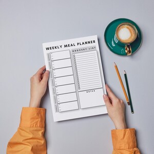 May include: A white weekly meal planner sheet with black text and lines, featuring a section for each day of the week, a grocery list with checkboxes, and a notes section. The planner is held against a plain background with a coffee cup and pencils.