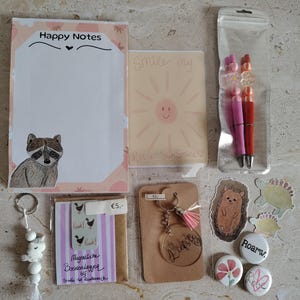 Op de afbeelding: Een briefpapier set met een notitieblok met een wasbeerillustratie en de tekst Happy Notes, een kaart met een zonnetje en de tekst Smile my little sunshine, twee pennen, een kralen sleutelhanger, een magnetische boekenlegger en diverse stickers.