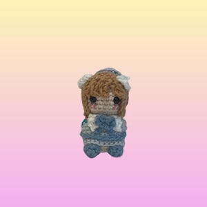 May include: A small crocheted doll featuring light brown yarn hair, two black bead eyes, and pink stitched cheeks. The doll wears a light blue and white dress with a blue bow detail at the centre, and a matching light blue hat with white accents.