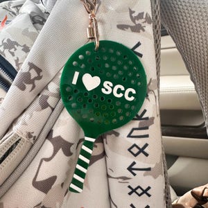 May include: A green circular paddle keychain with a perforated surface and a white handle featuring horizontal stripes. The center of the paddle displays the white text I heart SCC, where the heart is a solid white shape, attached by a metal clasp.