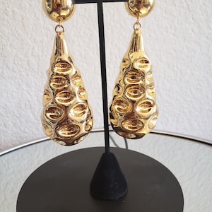 May include: A pair of gold-toned drop earrings featuring a smooth, spherical top stud connected to a large, elongated, teardrop-shaped pendant. The pendant surface is highly reflective and textured with multiple recessed, oval-shaped indentations.