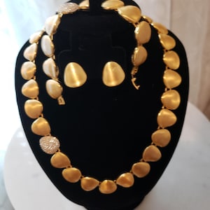 May include: A gold-toned jewelry set consisting of a necklace, a bracelet, and a pair of stud earrings. Each piece features smooth, matte-finish, teardrop-shaped links, with select links accented by a cluster of small, clear, sparkling crystals.