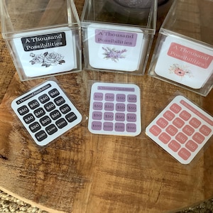 Könnte beinhalten: Drei durchsichtige, quadratische Acrylboxen, die jeweils eine Karte mit der Aufschrift A Thousand Possibilities und Blumenillustrationen enthalten. Neben jeder Box liegt eine passende quadratische Karte mit einem Raster aus sechzehn abgerundeten Quadraten, die mit $50 und $100 beschriftet sind.