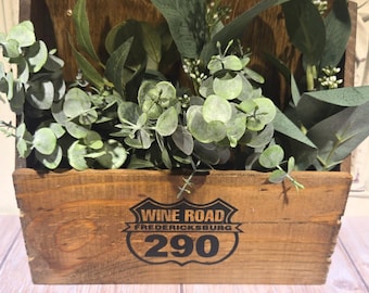 Puede incluir: Una caja rectangular de madera marrón llena de hojas de eucalipto artificiales verdes y pequeñas flores blancas. El frente de la caja presenta un estampado gráfico negro que dice WINE ROAD FREDERICKSBURG 290 en forma de escudo.