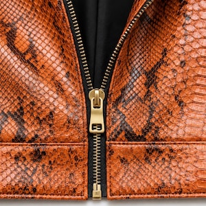 May include: A jacket featuring a textured orange and black snakeskin pattern. The garment has a central vertical zipper with a matte gold metal pull tab and a black fabric lining visible along the inner edges of the front opening.