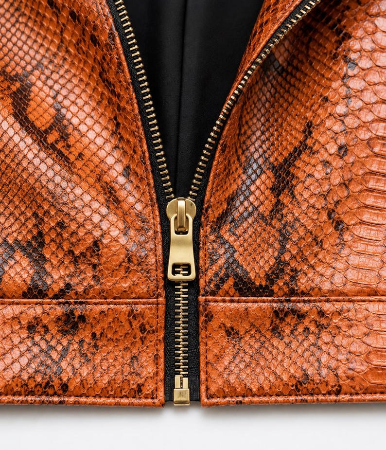 May include: A jacket featuring a textured orange and black snakeskin pattern. The garment has a central vertical zipper with a matte gold metal pull tab and a black fabric lining visible along the inner edges of the front opening.