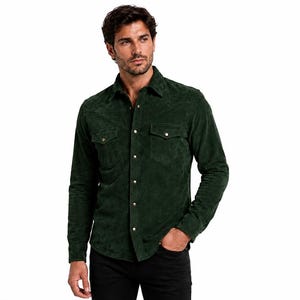 Puede incluir: Una camisa de manga larga de ante verde bosque oscuro con cuello en punta, dos bolsillos en el pecho con solapas y cierre frontal de botones con broches metálicos. La camisa tiene un acabado texturizado y se combina con pantalones negros.