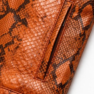 May include: A jacket featuring a textured orange and brown snakeskin pattern. The garment includes a vertical zippered pocket with a matching orange border and a horizontal seam detail along the bottom edge, showcasing a detailed scale-like surface.
