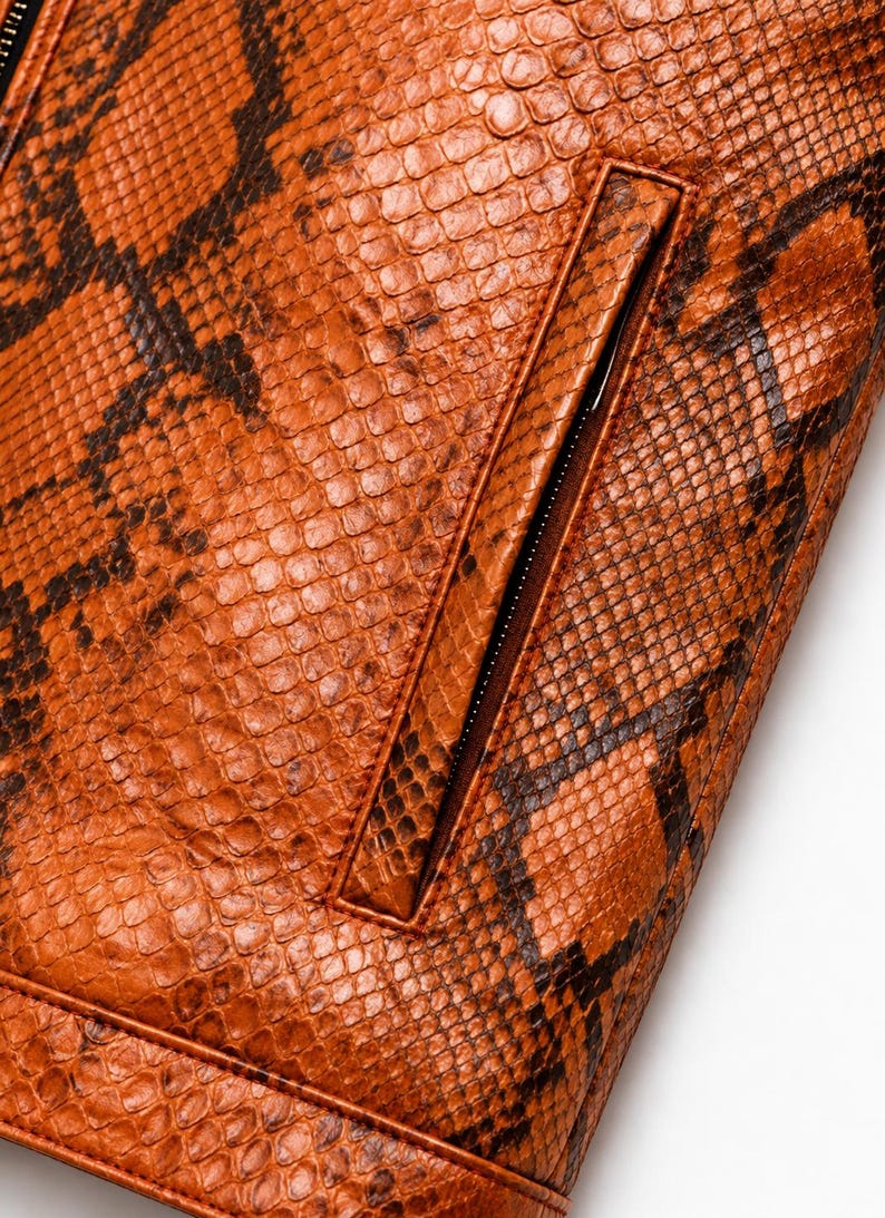 May include: A jacket featuring a textured orange and brown snakeskin pattern. The garment includes a vertical zippered pocket with a matching orange border and a horizontal seam detail along the bottom edge, showcasing a detailed scale-like surface.