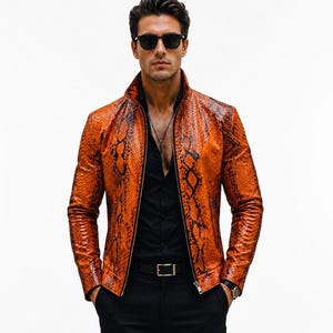 Puede incluir: Una chaqueta de cuero naranja y negra con estampado de piel de serpiente, cuello alto y cremallera frontal. La chaqueta se lleva sobre una camisa negra abotonada y pantalones negros, combinada con gafas de sol oscuras y un collar de plata.