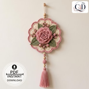 May include: A circular crochet wall hanging featuring a central dusty pink rose surrounded by green leaves and small berries on a cream-colored scalloped base. The piece is suspended by a cord with wooden beads and ends in a long dusty pink tassel.