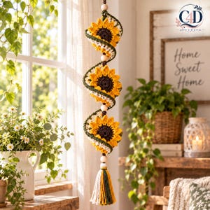 May include: A vertical hanging decoration featuring three crocheted sunflowers with dark brown centers and bright yellow petals, separated by wooden beads. The structure is wrapped in white and dark green yarn, ending in a multi-colored tassel.