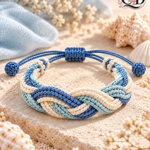 May include: A braided cord bracelet featuring a woven pattern of navy blue, light blue, and cream-colored threads. The bracelet includes an adjustable sliding knot closure with two small spherical cord ends in a matching navy blue color.