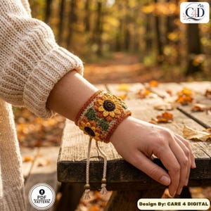 May include: A wide crochet wristband featuring a sunflower motif with yellow petals and dark brown centers on a textured background. The cuff is bordered in rust orange and includes two dangling beige cords with small wooden beads at the ends.